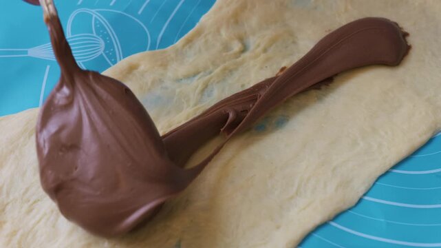 A thick line of chocolate hazelnut spread is piped onto a rectangular sheet of pale yellow dough. The dough sits on a blue silicone mat with measurement markings. Soft natural lighting.