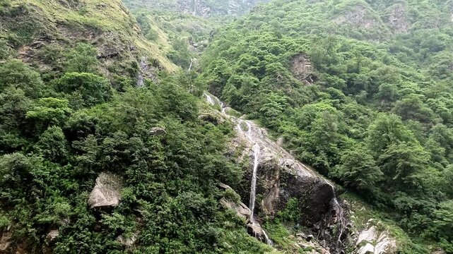 Natural waterfall flowing down rocky mountain slopes through a lush green Himalayan forest landscape. Suitable for nature, travel, monsoon, mountain scenery, forest, ecology, adventure tourism.