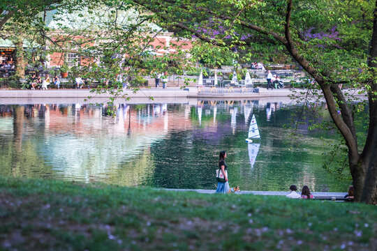 View of Central Park Conservatory Water with a model sailboat on the pond, people walking along the path, and lush green trees in spring New York, New York, United States.
