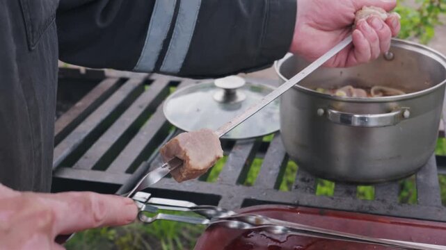Close-up of hands stringing marinated raw meat onto a metal skewer over an outdoor grill. Concept of summer recreation, outdoor cooking, and traditional barbecue preparation.