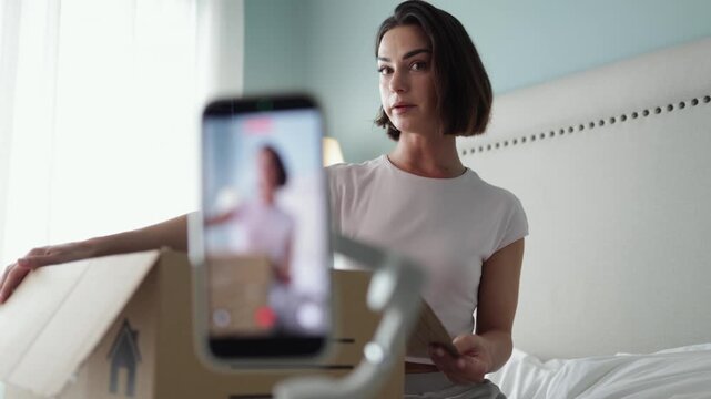 Content creator woman opening cardboard parcel and filming product review on mobile phone. Concept ecommerce, influencer lifestyle and retail.
