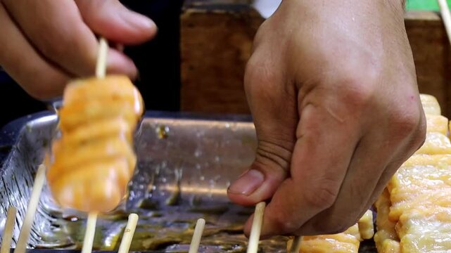 Close-up of a vendor using a teriyaki brush to apply a glaze or sauce to fresh salmon skewers at a street food market.