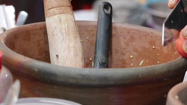 Close-up of a person slicing a tomato into a traditional clay mortar with a wooden pestle for Som Tum (Papaya Salad).
