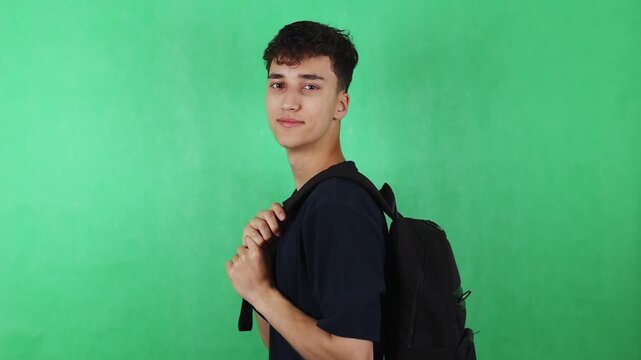 Young male student with dark hair adjusts backpack straps while smiling, standing against a bright green backdrop, showcasing a casual and relaxed demeanor in a studio setting