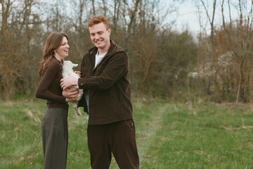 Naklejka premium Joyful couple outdoors holds a newborn goat kid, smiling at each other, casual attire, in a grassy field with trees, warm natural light, candid lifestyle moment showcasing rural charm and connection
