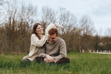 Naklejka premium Cheerful couple outdoors, close in with a playful goat kid by their side, cozy sweaters, sunlit meadow, natural colors, relaxed posture, candid moment, nature backdrop, lifestyle portrait