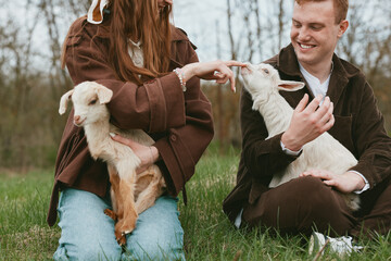 A couple with goats and kids share a tender outdoors moment in a grassy field, smiling and interacting with the animals, capturing a warm lifestyle scene of countryside companionship and farm life © SHOTPRIME STUDIO
