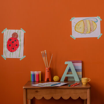 Creative kid corner with orange wall, children drawings, brushes, and colorful art supplies on a wooden desk. Bright and playful setup of room for learning and fun.