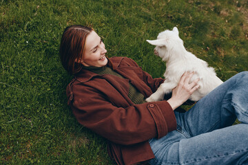 Naklejka premium Relaxed woman lying on grass wearing a brown jacket, holding a white goat kid, smiling while they share a gentle moment outdoors in a sunny meadow, natural lifestyle scene