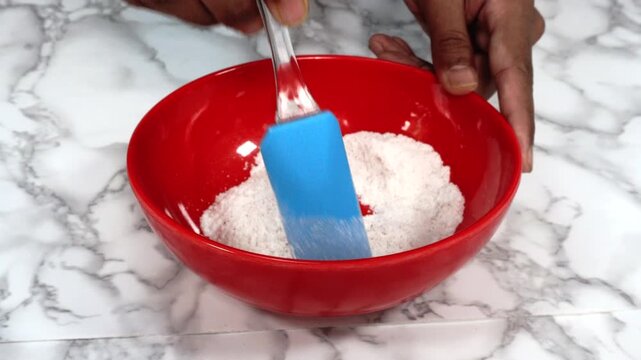 Stirring Dry Baking Ingredients in Red Bowl with Blue Spatula on Marble Countertop, Close-up of hands stirring dry baking ingredients in a bright red mixing bowl using a blue spatula