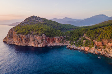 Obraz premium Aerial view of rocky Mediterranean coastline with forested hills and clear blue sea in Antalya region, Turkey. Coastal landscape at sunset with mountains and natural shoreline