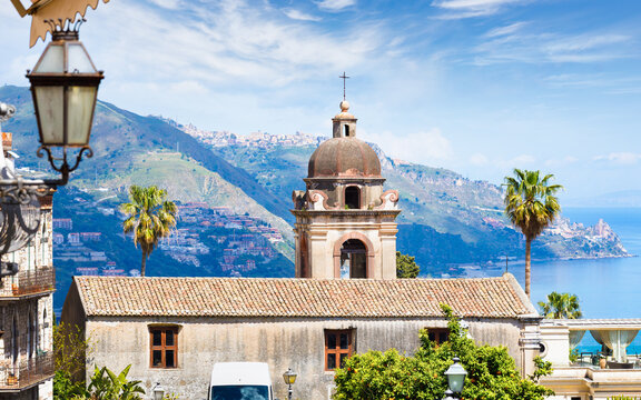 San Pancrazio Church above Mediterranean coast in Taormina, Sicily, Italy. Palm trees frame coastal landscape near Taormina with mountain villages under bright blue sky