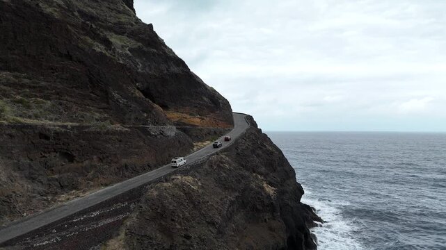 Aerial drone shot of a winding coastal road perched on the dark volcanic cliffs of Santo Antao, Cabo Verde. Vans travel along the narrow path overlooking the vast Atlantic Ocean at dusk