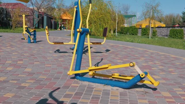 Outdoor fitness equipment for physical exercise stands on stone-paved ground in a sunny public park. The blue and yellow metal cross-trainer features steel handles with trees in background.