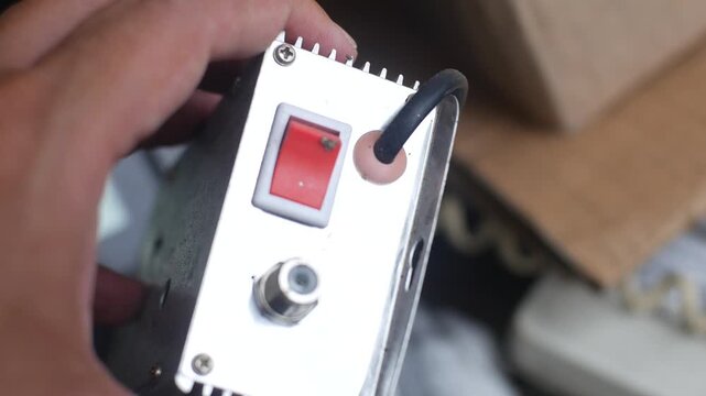A close-up video shows a hand holding and tilting a small silver electronic device equipped with a red power switch and various connectors, possibly a signal booster or radio transmitter.