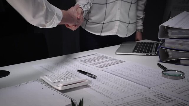 Business professionals shake hands over a cluttered desk of contracts, calculator and magnifying glass, sealing a trusted partnership after detailed financial review and late night work
