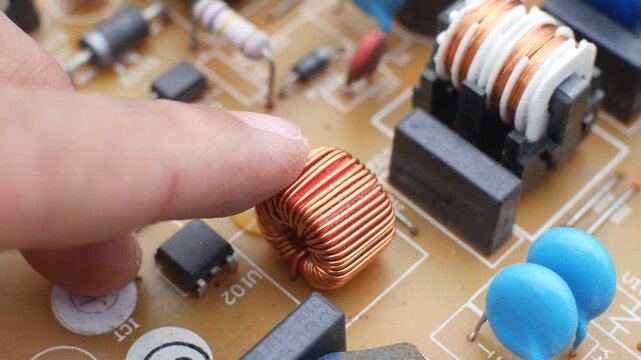 Close-up of a finger pointing at and touching a copper inductor coil on an electronic circuit board, surrounded by capacitors and other hardware components.