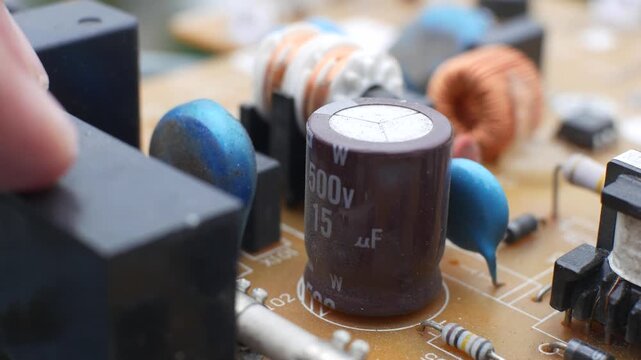 A close-up video captures a hand carefully inspecting various electronic components on a printed circuit board, focusing on a large electrolytic capacitor and its surrounding circuitry.