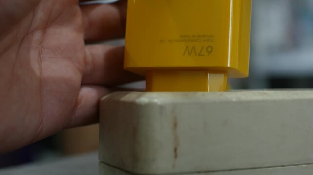 A person's hand connects a vibrant yellow 67W power adapter into a white electrical socket strip, demonstrating a simple everyday tech interaction and device setup.