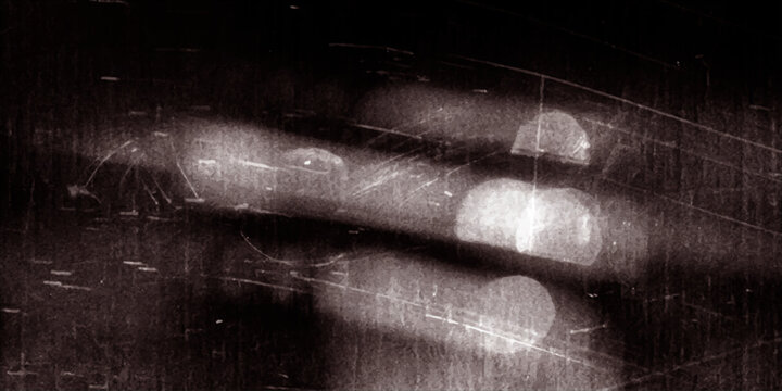The grunge texture of an old film projector lens reflects a cold winter night where white snow and rainy drops blur against the black metal of a wet car on a dark road