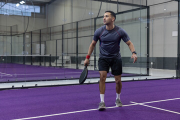 Fototapeta premium Adult man holding padel racket on purple-court with glass walls wearing grey top, red wristband