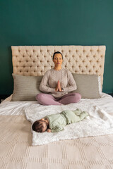 Obraz premium Mother sitting meditating with hands pressing on chest on bed wearing knit sweater, baby on blanket