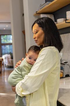 Cradling mother holding infant to chest in kitchen, wearing pale yellow shirt, light green onesie