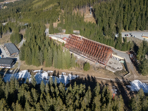 Aerial view of a large building under construction with a steel frame roof surrounded by dense pine forests and patches of melting snow Demanovska Dolina, Zilina Region, Slovakia.