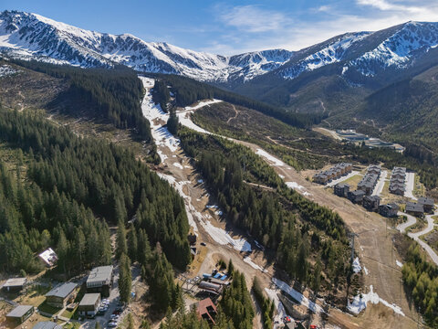 Aerial view of the Jasna ski resort area with melting snow on slopes, dense pine forests, and chalets nestled in the valley under the Low Tatras peaks in Demanovska Dolina, Zilina Region, Slovakia.