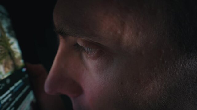 Close up profile of an adult man looking at a smartphone screen in a completely dark room. The bright digital display illuminates his facial features and reflects clearly in his eye.