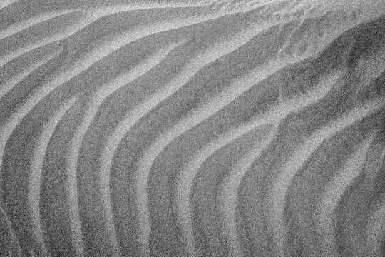 Black and white close-up photograph of wind-shaped sand dunes with soft ripples and natural patterns. The image highlights texture, curves, and minimal abstract forms, ideal for nature, desert, geolog