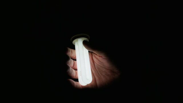 Hand holds an illuminated compact fluorescent lamp against a solid dark black background. Fingers safely touch the cool glass tubes. Energy saving and efficient electricity concept.