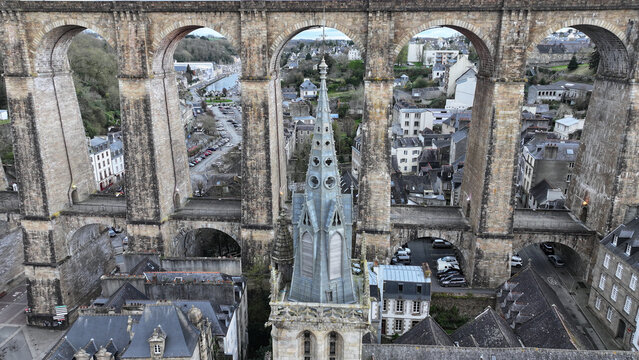 Aerial view of the Eglise Saint-Melaine spire framed by the massive stone arches of the historic railway viaduct and town rooftops in Morlaix, Bretagne, France.