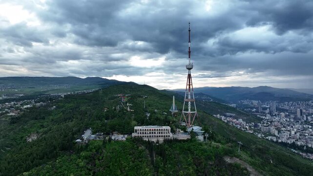 Aerial view of the Funicular Restaurant at Mtatsminda Park during sunset. Dynamic drone shot of a TV tower with Tbilisi city in the background.