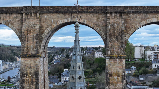 Aerial view of the spire of Eglise Saint-Melaine framed by the stone arches of the Morlaix Viaduct with the town and harbor in the background Morlaix, Bretagne, France.