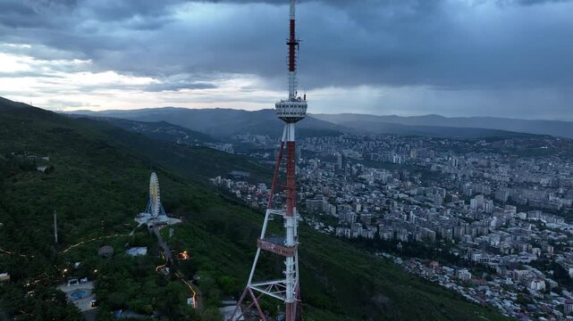 Aerial view of the Funicular Restaurant at Mtatsminda Park during sunset. Dynamic drone shot of a TV tower with Tbilisi city in the background.