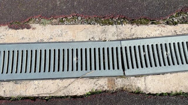 Galvanized drainage grate along stadium curb, long metal channel between asphalt and concrete, repeating slot pattern, leading