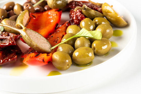 This antipasto plate features a colorful mix of green and Kalamata olives, fire-roasted red peppers, sun-dried tomatoes, and large caper berries drizzled with olive oil on a clean white background