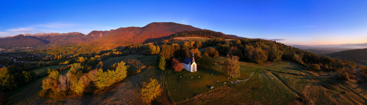 Aerial view of Chapelle Notre-Dame de Riantmont surrounded by autumn trees and rolling hills with mountains in the background during sunset Vesancy, Auvergne-Rhone-Alpes, France.