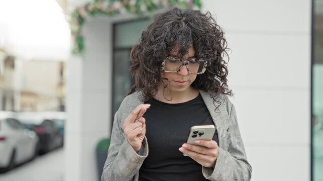 Young woman with curly hair and oversized glasses holding a smartphone, crossing fingers and reading a message while standing by a storefront on a street; anticipation hope.