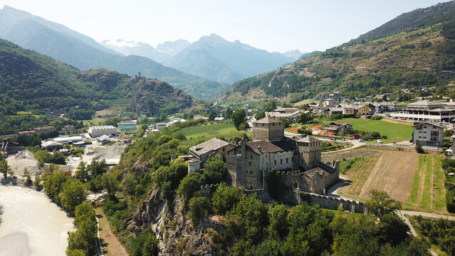 Aerial view of the Sarre Royal Castle on a rocky hill surrounded by lush greenery and a flowing river with the snow-capped Alps in the background in Aosta Valley, Aosta Valley, Italy.
