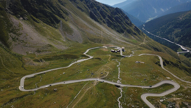 Aerial view of winding mountain road with hairpin turns crossing green alpine slopes and rocky peaks under a clear sky Aosta Valley, Italy.