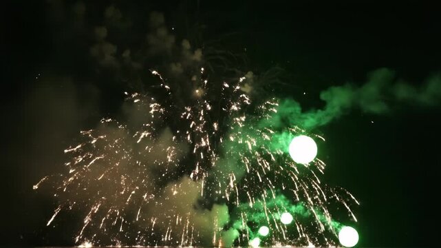 Several bright green and white firework bursts exploding simultaneously in a dark night sky with thick plumes of illuminated green smoke during a festive pyrotechnic display event.