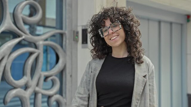 Woman waving hand by decorative metal gate at a building entrance, wearing glasses and checked blazer; friendly greeting.