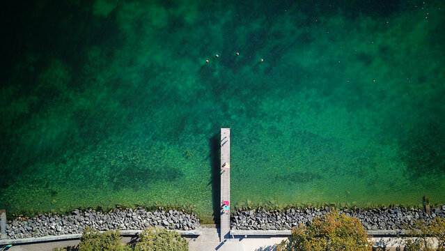Aerial view of a wooden pier extending into the turquoise waters of Lake Geneva with people standing on the deck in Saint-Prex, Vaud, Switzerland.