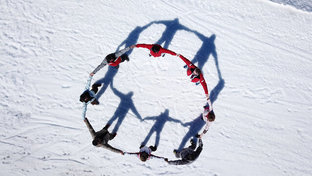 Aerial view of a group of people in winter gear holding hands in a circle on snow with long shadows cast on the white ground in Verbier, Val de Bagnes, Valais, Switzerland.