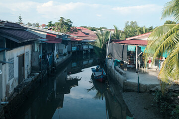 Obraz premium Quiet canal winds between modest houses. Red-tiled roofs reflect on calm water's surface. Palm fronds sway gently near a shaded doorway. Small boat rests near the canal's edge