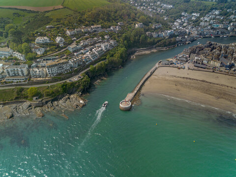 Aerial view of a boat travelling through the harbour entrance at Looe, Cornwall, beside Banjo Pier. The image shows turquoise coastal waters, sandy beach and the surrounding seaside town.