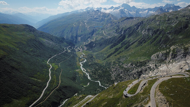 Aerial view of Furka Pass winding through a deep alpine valley with steep green slopes and snow-capped peaks in Obergoms, Valais, Switzerland.