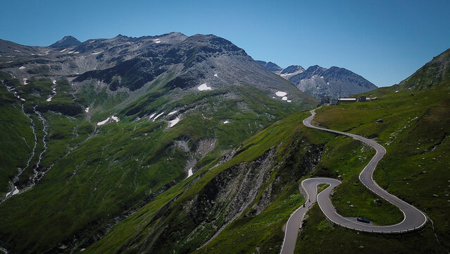 Aerial view of the winding hairpin turns of the Furka Pass mountain road surrounded by green alpine slopes and rocky peaks in Obergoms, Valais, Switzerland.